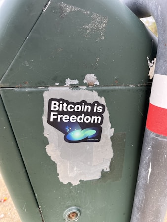 A sticker is placed on a green metal surface next to a metal pole. The sticker features the phrase 'Bitcoin is Freedom' with a graphic of abstract shapes, including a blue-green swoosh and small stars.
