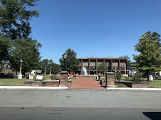 A well-maintained public square with a red brick path leads to a fountain in front of a two-story building labeled 'Union Bank.' The area is surrounded by trees and neatly trimmed lawns, with several street lamps and benches. A clear blue sky enhances the peaceful setting.