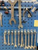 A collection of various hand tools organized neatly on a blue pegboard. Large wrenches are hanging at the top, alongside a screwdriver and pliers. Below are several smaller wrenches lined up in order of size. The pegboard also features a magnetic strip labeled for tool storage.