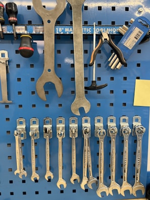 A collection of various hand tools organized neatly on a blue pegboard. Large wrenches are hanging at the top, alongside a screwdriver and pliers. Below are several smaller wrenches lined up in order of size. The pegboard also features a magnetic strip labeled for tool storage.