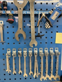 A collection of various hand tools organized neatly on a blue pegboard. Large wrenches are hanging at the top, alongside a screwdriver and pliers. Below are several smaller wrenches lined up in order of size. The pegboard also features a magnetic strip labeled for tool storage.
