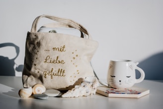 A small fabric bag with golden lettering stands next to a cute cat-faced mug. The bag features the words '...mit Liebe gefüllt' which translates to 'filled with love.' Near the bag, a collection of seashells and pebbles adds a natural element. The cat mug rests on a colorful coaster with a whimsical design, all placed on a light surface with soft shadows.