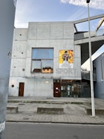 A modern concrete building with a minimalistic design, featuring a large window on the second floor and a poster of a boxer on the exterior wall. The word 'SPORT' is embedded in the concrete wall. The building entrance has glass doors and a bicycle rack is visible on the sidewalk. A streetlight and street are in the foreground, with a clear sky above.