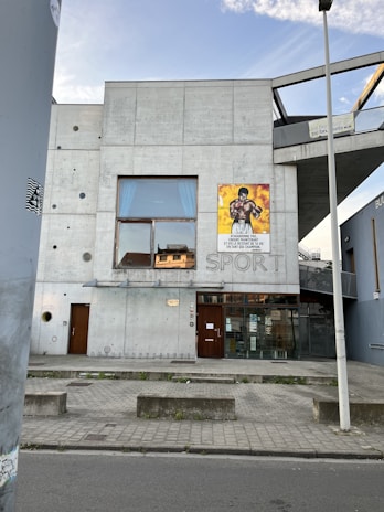 A modern concrete building with a minimalistic design, featuring a large window on the second floor and a poster of a boxer on the exterior wall. The word 'SPORT' is embedded in the concrete wall. The building entrance has glass doors and a bicycle rack is visible on the sidewalk. A streetlight and street are in the foreground, with a clear sky above.