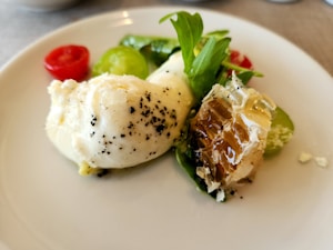 A plate featuring a piece of soft white cheese, possibly mozzarella, garnished with black pepper and accompanied by a chunk of honeycomb. Fresh arugula leaves and slices of red and green tomatoes add color, placed on a white dish against a neutral background.