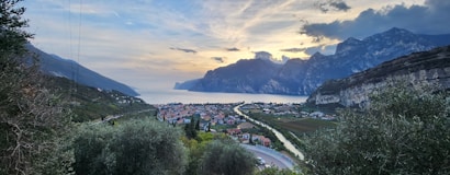 A picturesque landscape featuring a village nestled between lush greenery and a serene lake. Mountains tower in the background under a partly cloudy sky, with soft sunlight illuminating the scene. The village appears quiet and peaceful with houses, trees, and winding roads leading towards the lake.