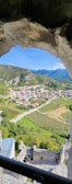 A picturesque view through a stone archway reveals a small town nestled in a lush valley. Rolling hills and mountains frame the background, while neatly arranged vineyards and fields spread across the landscape. Several buildings with red-tiled roofs are situated amidst the greenery, connected by winding roads.