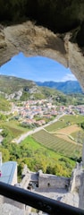 A picturesque view through a stone archway reveals a small town nestled in a lush valley. Rolling hills and mountains frame the background, while neatly arranged vineyards and fields spread across the landscape. Several buildings with red-tiled roofs are situated amidst the greenery, connected by winding roads.
