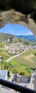 A picturesque view through a stone archway reveals a small town nestled in a lush valley. Rolling hills and mountains frame the background, while neatly arranged vineyards and fields spread across the landscape. Several buildings with red-tiled roofs are situated amidst the greenery, connected by winding roads.