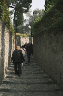 A group of people walking up a narrow stone pathway bordered by tall stone walls. The path is shaded by trees, and the atmosphere feels secluded and quiet. The pathway is lined with some greenery and an old-fashioned street lamp is visible. The scene suggests an uphill journey.