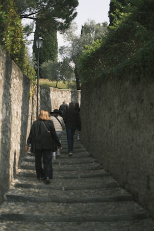A group of people walking up a narrow stone pathway bordered by tall stone walls. The path is shaded by trees, and the atmosphere feels secluded and quiet. The pathway is lined with some greenery and an old-fashioned street lamp is visible. The scene suggests an uphill journey.