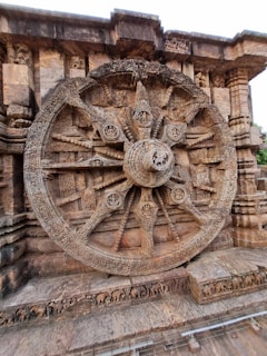 An intricately carved stone wheel is set against a temple wall, featuring detailed motifs and designs typical of ancient Indian architecture. The structure includes spokes and hub details with elaborate carvings and relief work.