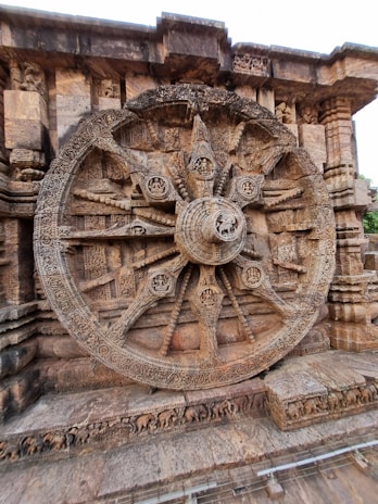 An intricately carved stone wheel is set against a temple wall, featuring detailed motifs and designs typical of ancient Indian architecture. The structure includes spokes and hub details with elaborate carvings and relief work.