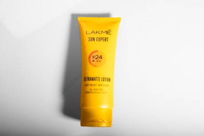 A yellow tube of Lakmé Sun Expert Ultramatte Lotion is placed against a plain white background. The product details indicate SPF 24 and PA++, highlighting its lightweight, non-sticky formula suitable for all skin types. The design is minimalistic, focusing on the brand and product benefits.