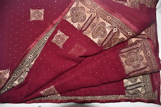 A richly patterned fabric featuring intricate gold designs on a deep maroon background. The designs include geometric and floral motifs, bordered with elaborate embroidery. The material appears luxurious and detailed.