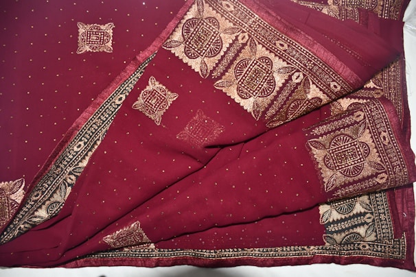 A richly patterned fabric featuring intricate gold designs on a deep maroon background. The designs include geometric and floral motifs, bordered with elaborate embroidery. The material appears luxurious and detailed.