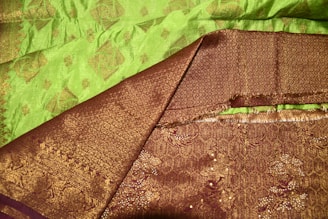 A rich, intricately patterned textile with a combination of green and deep bronze colors. The fabric features traditional motifs, with a smooth and shiny appearance, highlighting its luxurious texture.