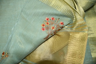 A close-up of an elegant fabric featuring intricate floral embroidery. The fabric consists of a blend of soft blue and golden tones, with detailed red flower designs woven into it. The texture appears rich and smooth, highlighting the fine craftsmanship.