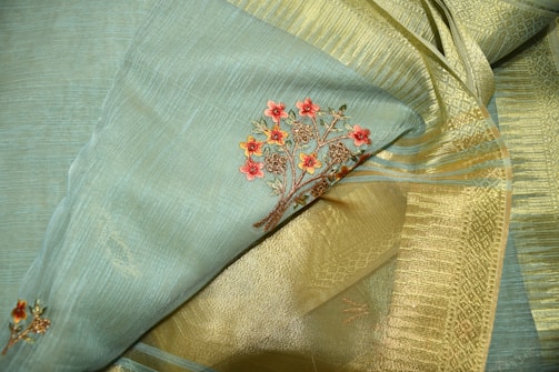 A close-up of an elegant fabric featuring intricate floral embroidery. The fabric consists of a blend of soft blue and golden tones, with detailed red flower designs woven into it. The texture appears rich and smooth, highlighting the fine craftsmanship.