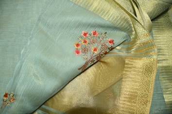 A close-up of an elegant fabric featuring intricate floral embroidery. The fabric consists of a blend of soft blue and golden tones, with detailed red flower designs woven into it. The texture appears rich and smooth, highlighting the fine craftsmanship.