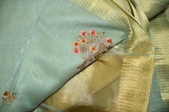 A close-up of an elegant fabric featuring intricate floral embroidery. The fabric consists of a blend of soft blue and golden tones, with detailed red flower designs woven into it. The texture appears rich and smooth, highlighting the fine craftsmanship.