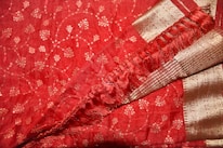A richly detailed red fabric with intricate gold embroidery forming small floral patterns. The fabric also features decorative gold borders with horizontal stripes and a section of delicate fringe, adding texture and elegance.