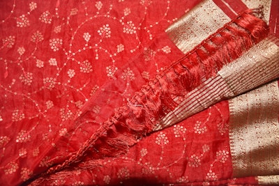 A richly detailed red fabric with intricate gold embroidery forming small floral patterns. The fabric also features decorative gold borders with horizontal stripes and a section of delicate fringe, adding texture and elegance.
