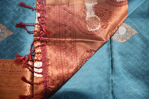 A richly woven fabric with intricate patterns and textures. The cloth features a combination of teal and gold colors, with a zigzag pattern on the teal section and ornate paisley designs on the gold section. A burgundy border with tassels adds elegance to the textile.