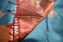 A richly woven fabric with intricate patterns and textures. The cloth features a combination of teal and gold colors, with a zigzag pattern on the teal section and ornate paisley designs on the gold section. A burgundy border with tassels adds elegance to the textile.