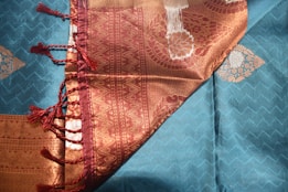 A richly woven fabric with intricate patterns and textures. The cloth features a combination of teal and gold colors, with a zigzag pattern on the teal section and ornate paisley designs on the gold section. A burgundy border with tassels adds elegance to the textile.