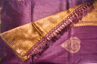 A richly woven textile featuring intricate designs. The fabric is primarily dark purple with golden embellishments, including a decorative border and detailed patterns. The golden area includes a series of interlocking shapes and fringe on one side, adding texture and elegance.