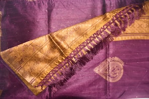 A richly woven textile featuring intricate designs. The fabric is primarily dark purple with golden embellishments, including a decorative border and detailed patterns. The golden area includes a series of interlocking shapes and fringe on one side, adding texture and elegance.