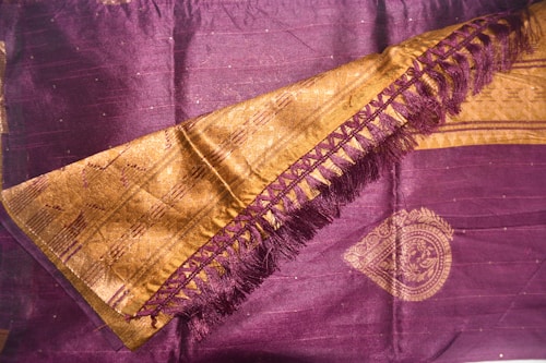 A richly woven textile featuring intricate designs. The fabric is primarily dark purple with golden embellishments, including a decorative border and detailed patterns. The golden area includes a series of interlocking shapes and fringe on one side, adding texture and elegance.