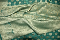 An elaborate teal fabric featuring intricate gold embroidery and patterns with floral and geometric designs. The texture appears rich and luxurious, indicating high-quality material. The fabric is draped elegantly, showcasing its sheen and craftsmanship.