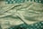 An elaborate teal fabric featuring intricate gold embroidery and patterns with floral and geometric designs. The texture appears rich and luxurious, indicating high-quality material. The fabric is draped elegantly, showcasing its sheen and craftsmanship.