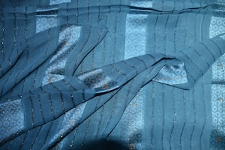 A fabric with a textured pattern featuring horizontal stripes and intricate designs, highlighted with subtle metallic threads. The textile appears to be made of a soft material with a smooth, flowing drape.