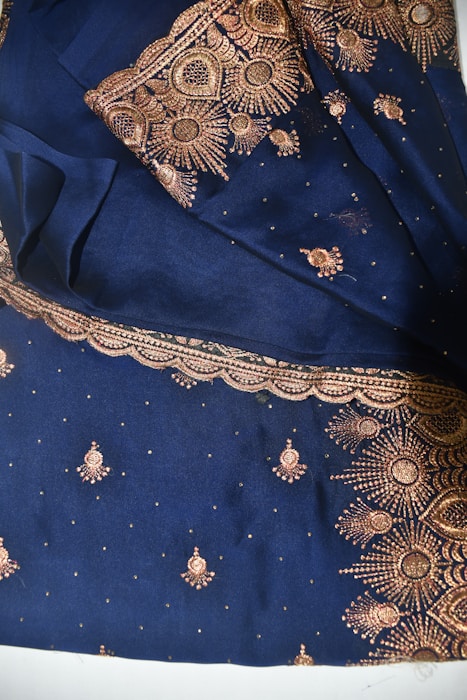 A rich blue fabric adorned with intricate gold embroidery, featuring floral and paisley patterns. The ornate designs are meticulously stitched with glittering gold thread, highlighting an elaborate and traditional textile craftsmanship.