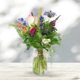 A vibrant bouquet of mixed seasonal flowers in soft pastel tones, arranged elegantly in a clear glass vase.