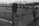 A monochrome image featuring a close-up view of a wooden barrier wrapped with barbed wire. In the background, there is a road with multiple lanes and a metal railing, possibly near a coastal area as there’s an indication of water in the distance.