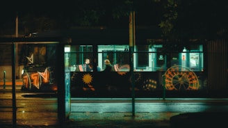 A nighttime street scene featuring a bus with illuminated, colorful graffiti designs including roulette and dice motifs. Inside the bus, two people are sitting across from each other, visible through the large windows. A dimly lit bus stop with reflective surfaces and wet pavement glistening from the streetlights adds to the ambiance.
