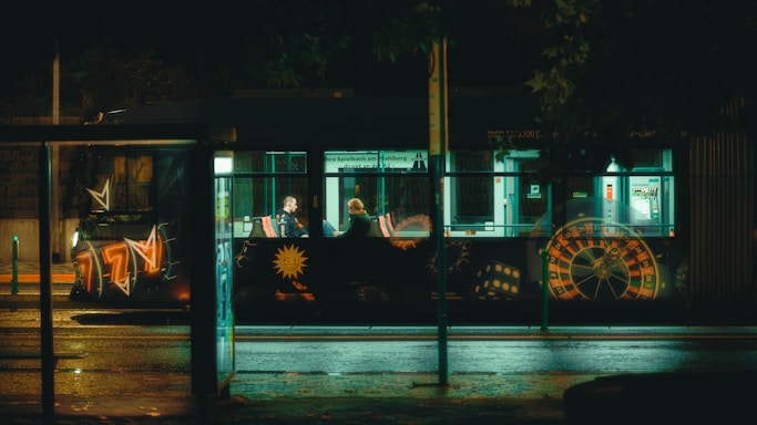 A nighttime street scene featuring a bus with illuminated, colorful graffiti designs including roulette and dice motifs. Inside the bus, two people are sitting across from each other, visible through the large windows. A dimly lit bus stop with reflective surfaces and wet pavement glistening from the streetlights adds to the ambiance.