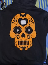A black hoodie features an orange skull design in the style of a sugar skull with floral and heart embellishments. Positioned at the center of the design is a white charging case for wireless earbuds, with each earbud placed in the eyes of the skull. The hoodie is laid out on a background of striped and solid-colored fabrics.
