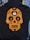 A black hoodie features an orange skull design in the style of a sugar skull with floral and heart embellishments. Positioned at the center of the design is a white charging case for wireless earbuds, with each earbud placed in the eyes of the skull. The hoodie is laid out on a background of striped and solid-colored fabrics.