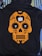 A black hoodie features an orange skull design in the style of a sugar skull with floral and heart embellishments. Positioned at the center of the design is a white charging case for wireless earbuds, with each earbud placed in the eyes of the skull. The hoodie is laid out on a background of striped and solid-colored fabrics.