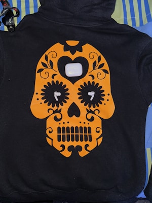 A black hoodie features an orange skull design in the style of a sugar skull with floral and heart embellishments. Positioned at the center of the design is a white charging case for wireless earbuds, with each earbud placed in the eyes of the skull. The hoodie is laid out on a background of striped and solid-colored fabrics.