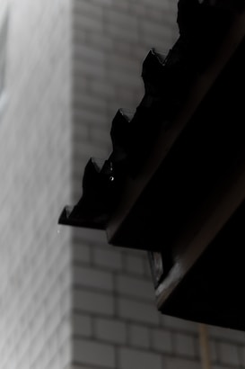 A close-up of a dark, corrugated roof edge with a blurred brick wall in the background. Water droplets can be seen hanging from the roof, indicating damp or rainy conditions.