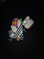 A pair of white wireless earbuds is placed on top of a graphic design. The design features stylized white stripes surrounded by colorful flowers and green leaves, set against a dark background.
