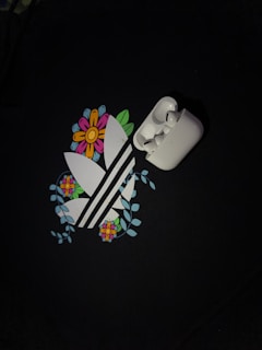A pair of white wireless earbuds is placed on top of a graphic design. The design features stylized white stripes surrounded by colorful flowers and green leaves, set against a dark background.