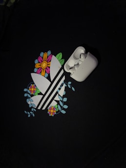 A pair of white wireless earbuds is placed on top of a graphic design. The design features stylized white stripes surrounded by colorful flowers and green leaves, set against a dark background.