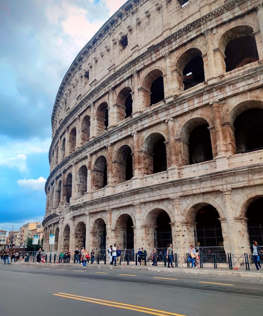 The Colosseum, an ancient Roman amphitheater, is partially visible, showcasing its iconic arches and stone construction. A group of tourists and visitors are gathered around, indicating its popularity as a historic site. The sky is partly cloudy, providing a dramatic backdrop to the robust structure.